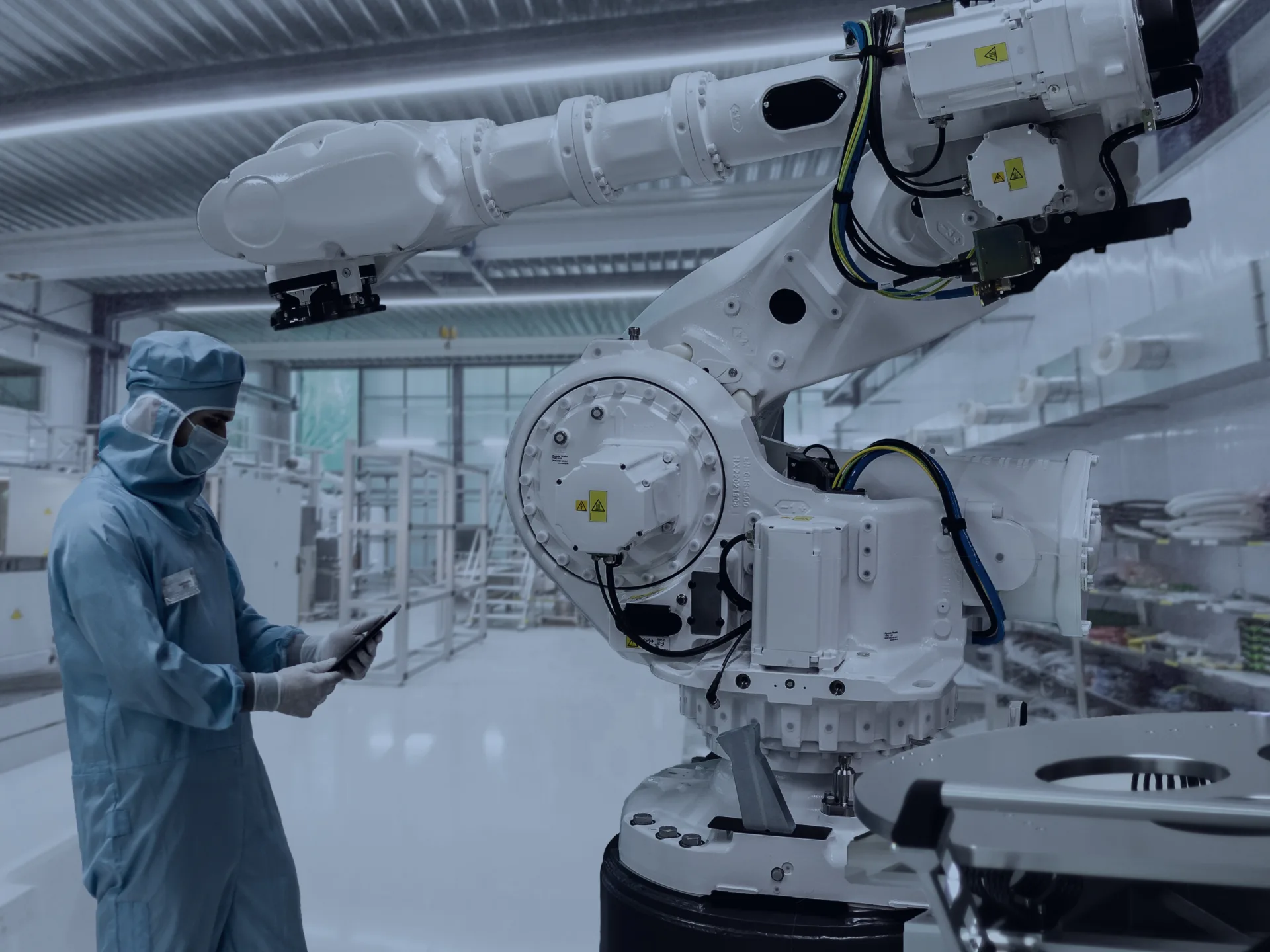 Temiz odada beyaz altı eksenli endüstriyel robot kolu ve tablete bakan tulumlu, maskeli operatör
